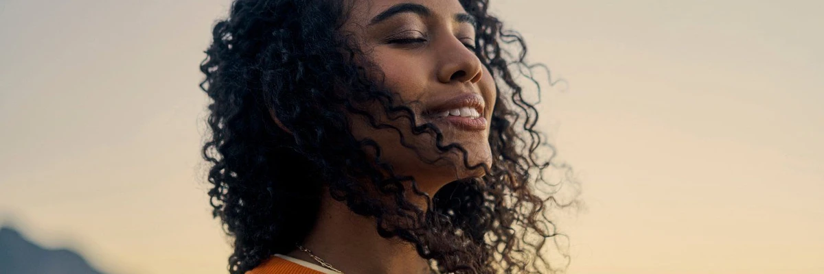Femme aux cheveux bouclés souriant les yeux fermés sous un ciel clair.