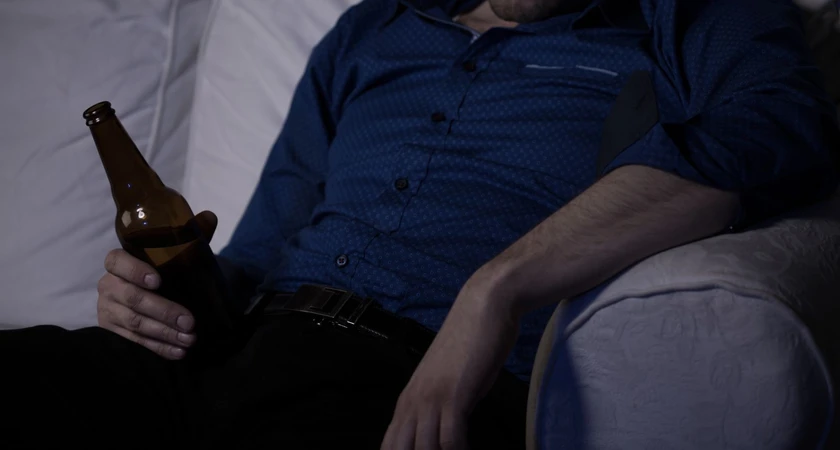 Homme assis sur un canapé, portant une chemise bleue, tenant une bouteille de bière.
