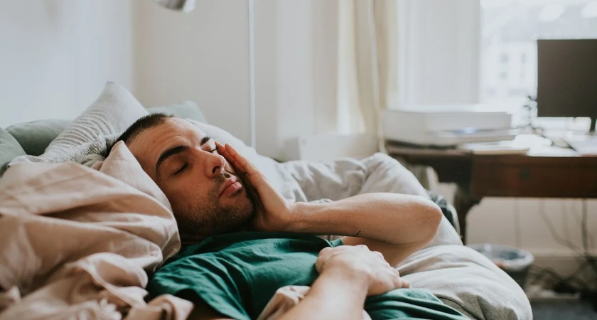 Homme allongé sur un lit, semblant somnolent, main sur le visage dans une chambre lumineuse.
