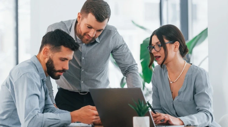two men and a woman in an office look at some information on a laptob