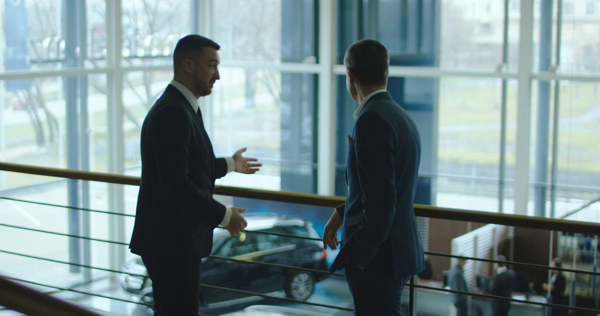 Two men having conversation on balcony in car showroom.
