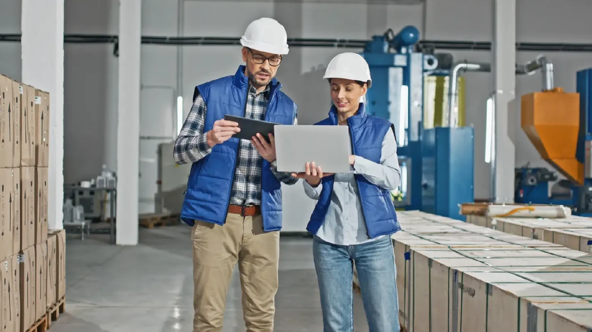 Two Caucasian workers wearing uniforms and helmets while standing in large storage or industrial facility. 