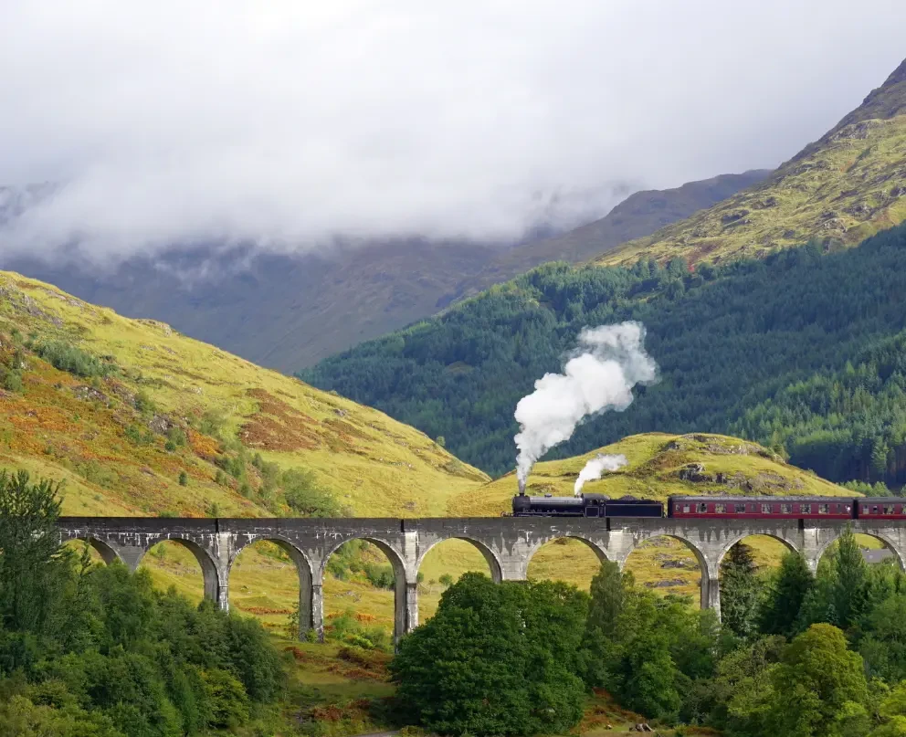 A steam train running along a mountianous track wih vibrant trees in the background