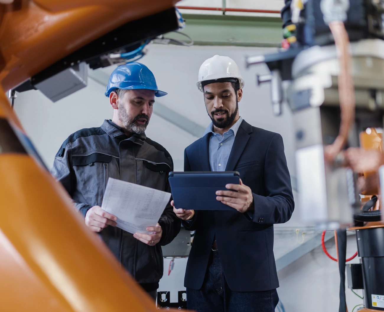 Two professional men working on tablet looking at software that helps optimize work in their industrial manufacturing plant. Big machines are seen in the background.