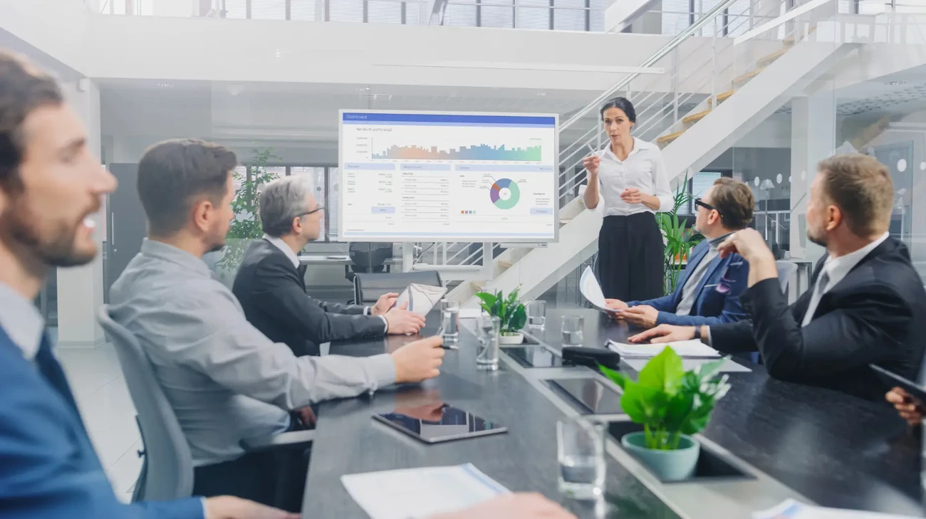 A dynamic team gathered around a meeting table, where a woman is explaining some information and infographs on a board to the team members