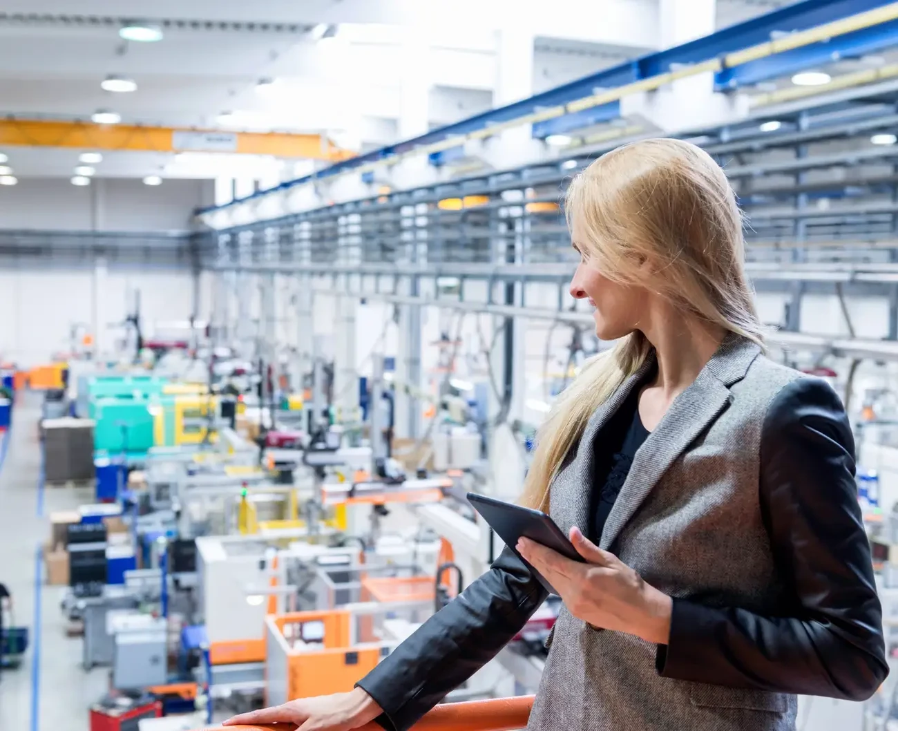 A business woman is standing on a balcony overlooking a manufacturing floor and monitoring the digital transformation of the factory on a tablet
