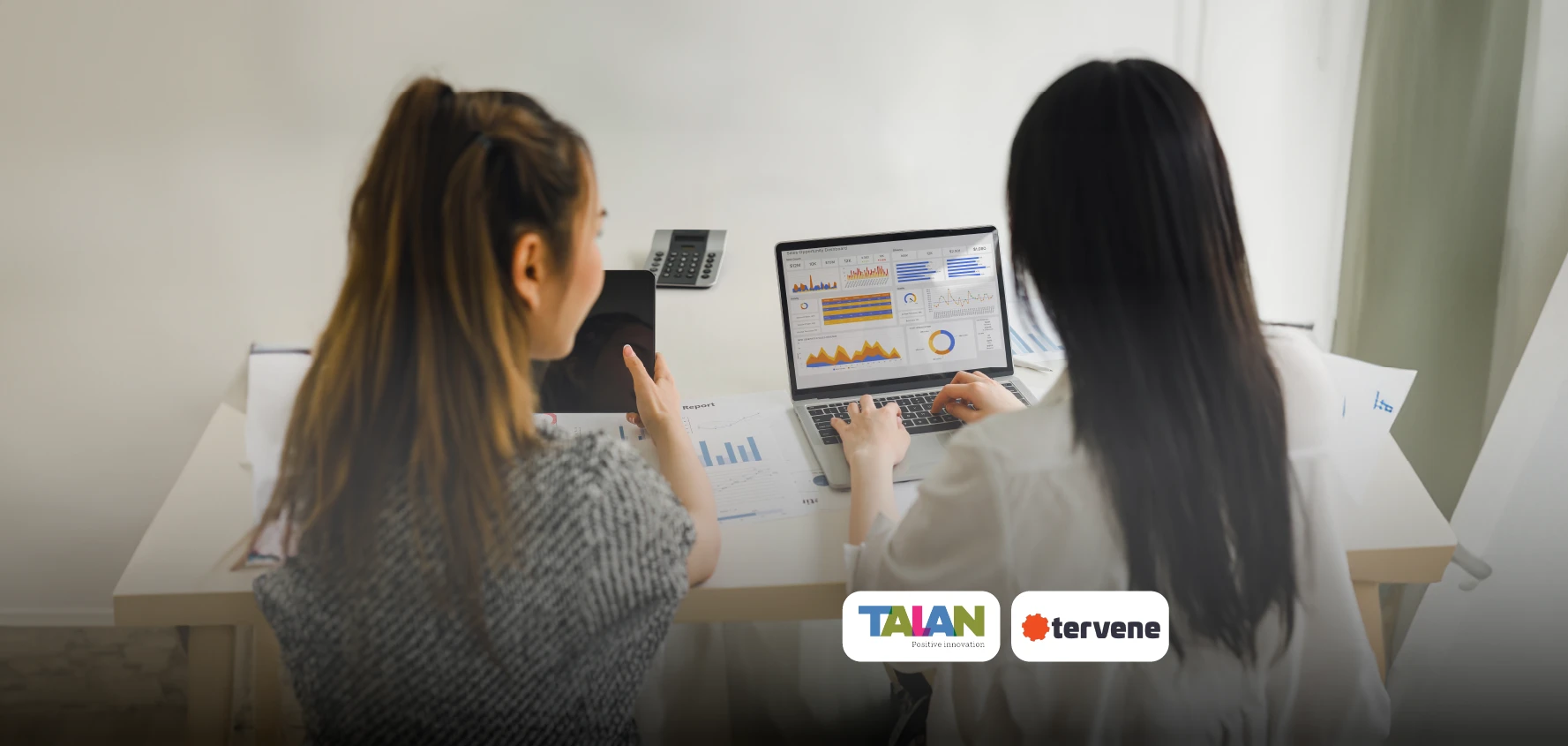 Two female co-workers specializing in DMS (Daily Management System) with Tervene. They are reviewing the daily management software designed for the manufacturing industry on a computer and a tablet.