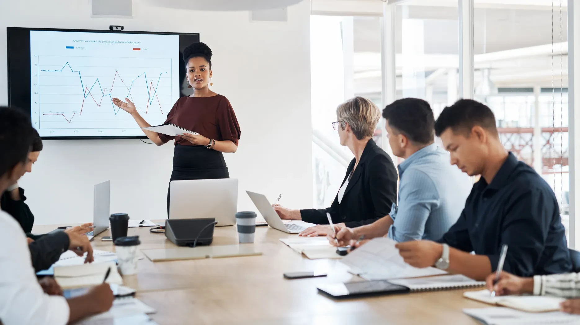 A dynamic team gathered around a desk at work, where a woman is explaining some relevant data and inforgarphs on a borad to the other team members
