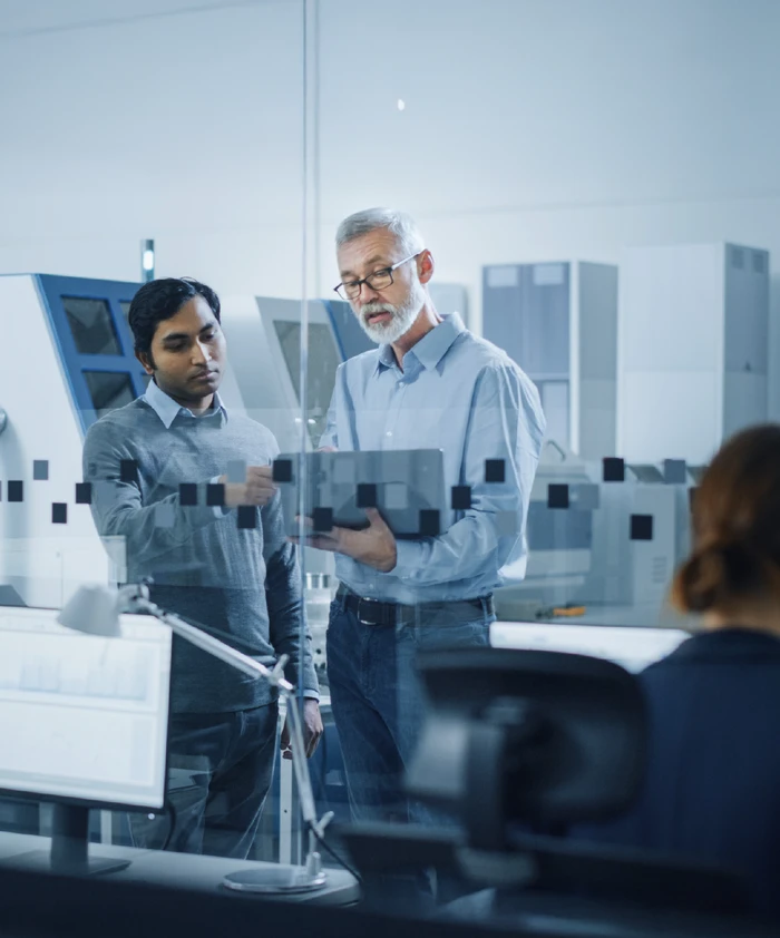 Two engineers looking at the sap software computer in an office full of large machines in a manufacturing plant. 