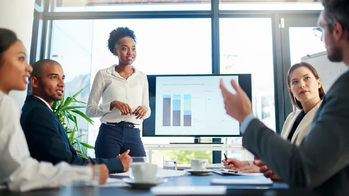 Female leader analyzing data in front of a team of potential clients. Digital screen and computers are seen.