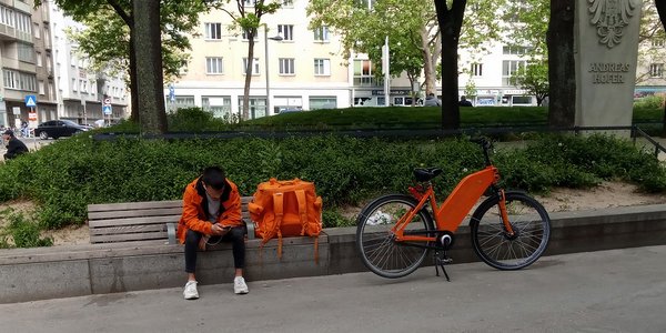 Eine Person in orangefarbener Kleidung sitzt auf einer Bank und schaut auf ihr Handy. Neben ihr steht ein orangefarbenes Fahrrad und eine große Umhängetasche. Im Hintergrund sieht man Bäume und Gebäude, die für eine städtische Umgebung typisch sind.