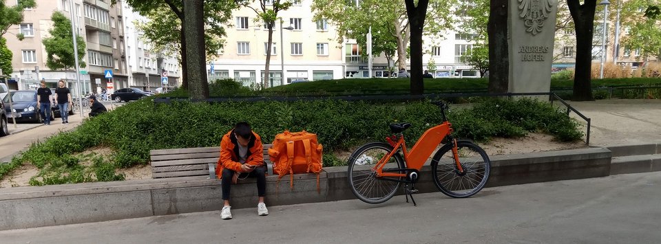 Eine Person in orangefarbener Kleidung sitzt auf einer Bank und schaut auf ihr Handy. Neben ihr steht ein orangefarbenes Fahrrad und eine große Umhängetasche. Im Hintergrund sieht man Bäume und Gebäude, die für eine städtische Umgebung typisch sind.
