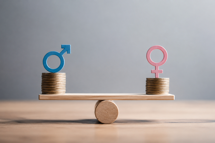 Die Abbildung zeigt eine Wippe mit zwei Stapeln Münzen, auf denen Symbole für Geschlechter platziert sind: ein blaues Männer-Symbol a.d. linken Seite und ein rosa Frauen-Symbol auf der rechten Seite. Das Bild thematisiert die Gleichheit der Geschlechter.