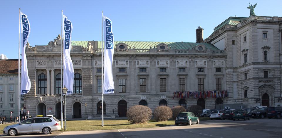 Das Bild zeigt ein historisches Gebäude in Wien, das die Flaggen der OSZE an den Fahnenmasten schwingt. Im Vordergrund parken einige Autos, während im Hintergrund eine grüne Rasenfläche und weitere Bäume sichtbar sind. Der Himmel ist blau und klar.