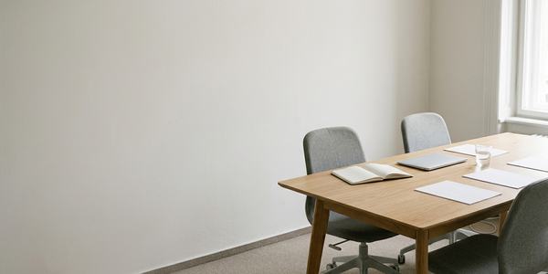 Ein heller Besprechungsraum mit einem Holz-Tisch und grauen Stühlen. Auf dem Tisch liegen ein Notizbuch, ein Laptop und einige Blätter. Die Wände sind schlicht und weiß, und ein Fenster lässt viel Tageslicht herein.