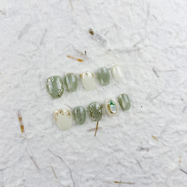 Ongles courts ovales frais à presser en vert pâle et blanc avec des fleurs blanches, des touches dorées et une finition brillante, parfaits pour les