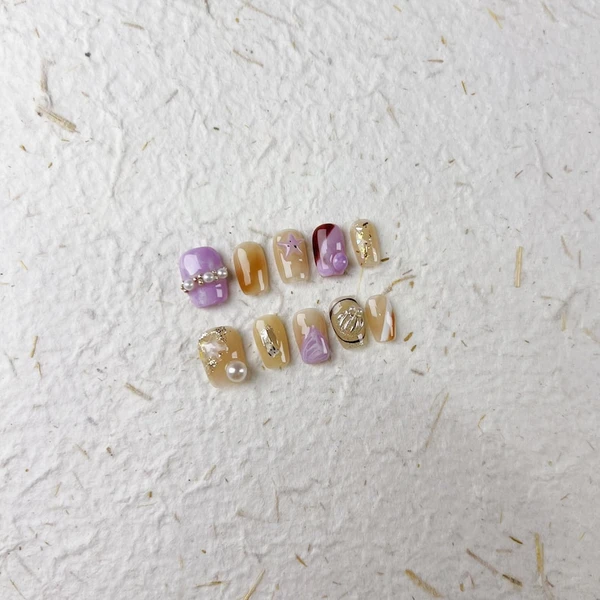 Ongles à presser carrés courts brillants en violet, pêche avec des chaînes de perles, des étoiles de mer et des touches de feuille d'or. Un style
