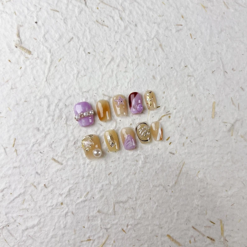 Ongles à presser carrés courts brillants en violet, pêche avec des chaînes de perles, des étoiles de mer et des touches de feuille d'or. Un style