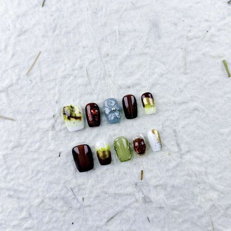 Ongles courts et vifs à presser dans des tons de blanc moucheté doré, de vin rouge foncé, de fleurs bleu clair et de brun à or pour un look artistique