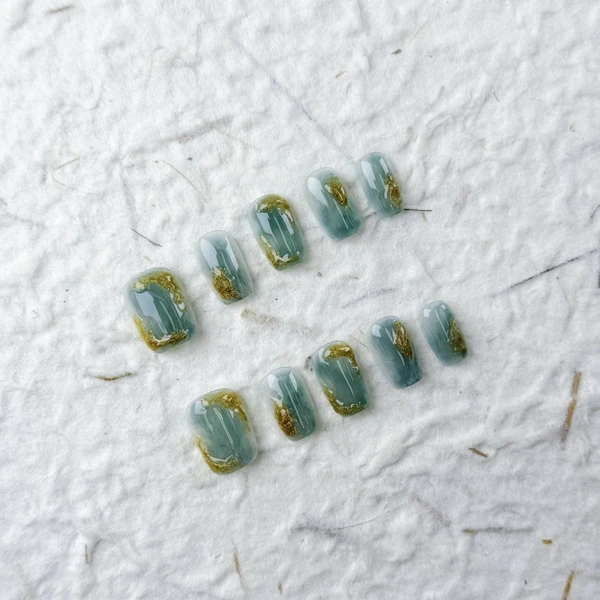 Presse à ongles carrée artistique courte en gris-vert brumeux, avec des textures dorées comme des ondulations de lac et une finition brillante,